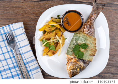 Top view of baked dorado fish in bamboo leaves and Szechuan sauce, baked potatoes with bamboo shoots on a white plate. 110593371