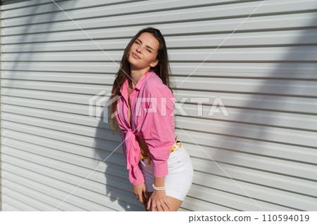 Young beautiful brunette woman in a pink shirt and white denim shorts against the background of a light garage door fence 110594019