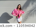 Young beautiful brunette woman in a pink shirt and white denim shorts against the background of a light garage door fence 110594042