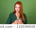 Young red hair woman on green background with mobile phone texting chatting angry disappointed serious 110594048
