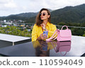 Stylish fit fashion women in bright yellow shirt trendy sunglasses posing at rooftop terrace tropical view outdoor holding mobile phone 110594049
