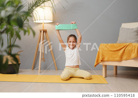 Smiling cheerful kid girl using fitness rubber resistance band exercising at home having workout in living room training her arms doing physical exercises with good mood enjoying healthy lifestyle 110595418