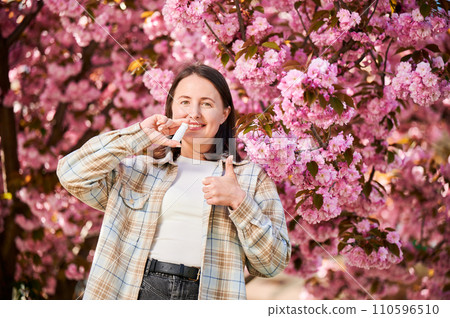 Woman allergic using medical nasal spray, suffering from seasonal allergy at spring in blossoming garden. Young woman treating runny nose in front of blooming tree outdoors. Spring allergy concept. 110596510