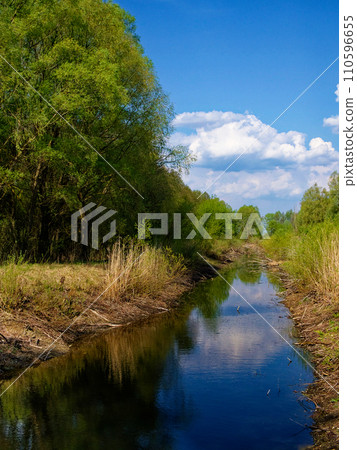 Clear skies and fluffy clouds reflect in a tranquil, tree-lined waterway. Clear skies and fluffy clouds reflect in a tranquil, tree-lined waterway. 110596655