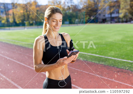 Sporty female athlete in fitness attire enjoying music while interacting with her phone on a sunny track field. Sporty female athlete in fitness attire enjoying music while interacting with her phone on a sunny track field. 110597292