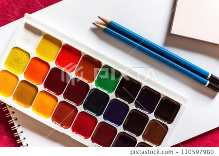 Colorful watercolor or acrylic paints and pencils on a white sheet of paper on red background. Design studio table top view. Colorful watercolor or acrylic paints and pencils on a white sheet of paper on red background. Design studio table top view. 110597980