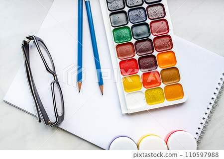 Colorful watercolor or acrylic paints, pencils and glasses on a white sheet of paper on a white background. Design studio table top view.  110597988