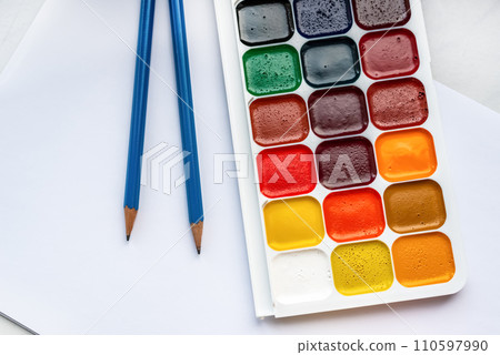 Colorful watercolor or acrylic paints and pencils on a white sheet of paper on a white background. Design studio table top view. Colorful watercolor or acrylic paints and pencils on a white sheet of paper on a white background. Design studio table top view. 110597990