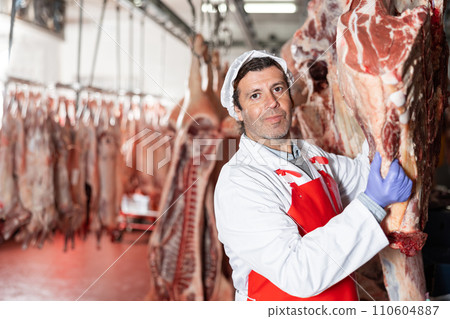 Male slaughterhouse worker showing beef chunk in meat storage 110604887