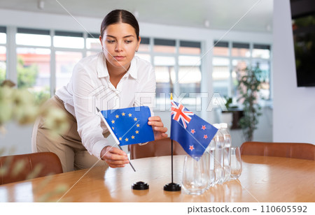 Young woman puts EU and New Zealand flags on table in office 110605592