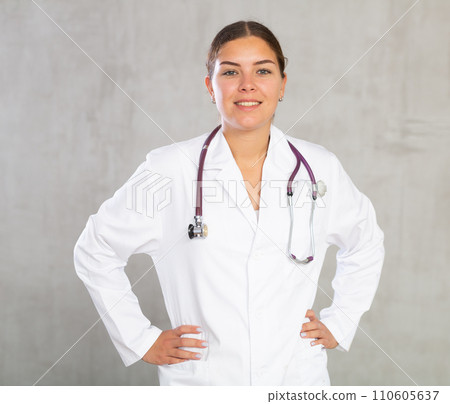 Friendly female professional doctor in white coat posing against gray studio background, looking at camera with smile Friendly female professional doctor in white coat posing against gray studio background, looking at camera with smile 110605637