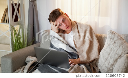 Stressful work. Tired freelancer. Remote sick employee man suffering from headache sitting on couch in blanket with laptop at home in living room. 110608742