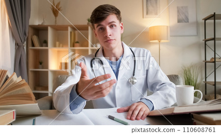 Doctor consultation. Medical support. Telehealth help. Specialist man in white hospital uniform explaining sitting at work desk at video appointment in light clinic interior. 110608761