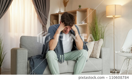 Headache suffer. Sickness symptoms. Exhausted man grasping head feeling bad covered with plaid sitting on couch in living room home interior. Headache suffer. Sickness symptoms. Exhausted man grasping head feeling bad covered with plaid sitting on couch in living room home interior. 110608784