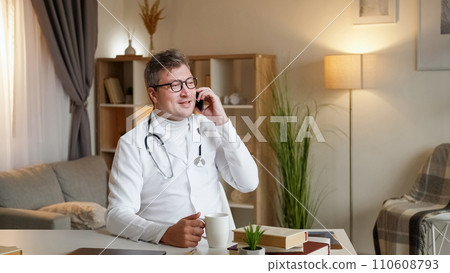 Doctor phone talk. Mobile call. Work break. Positive hospital specialist man enjoying cell communication free time drinking coffee sitting at clinic desk. 110608793