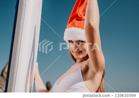 Woman sea sup. Close up portrait of happy young caucasian woman with long hair in Santa hat looking at camera and smiling. Cute woman portrait in a white bikini posing on sup board in the sea Woman sea sup. Close up portrait of happy young caucasian woman with long hair in Santa hat looking at camera and smiling. Cute woman portrait in a white bikini posing on sup board in the sea 110610309