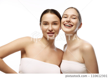 Portrait of two women with natural makeup, highlighting their clear skin and subtle beauty features looking at camera against white background. 110612301