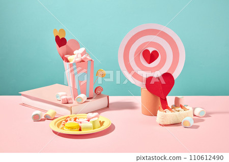 A dish containing gummy candy, marshmallow and macaroon. A paper bucket filled with candies placed on a book.Paper bullseye with red heart displayed on a ;podium. Happy Valentine day 110612490