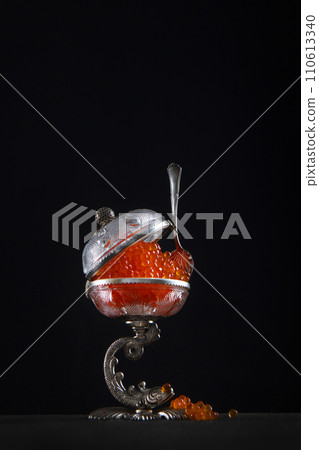 Red caviar. Caviar in the caviar bowl. A spoon for caviar. Beautiful vintage tableware. Black background. Vertical photo. Copy space. 110613340