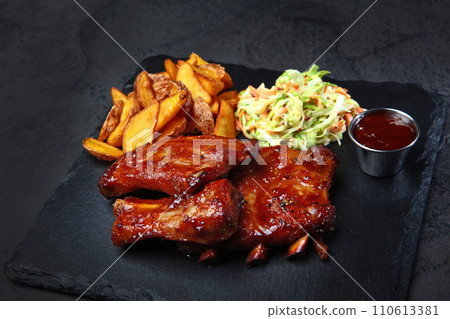 Barbecue pork ribs with baked potatoes and fresh cabbage salad on a black background. Barbecue pork ribs with baked potatoes and fresh cabbage salad on a black background. 110613381