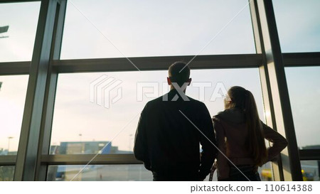 Attractive young woman and handsome man with suitcases are ready for traveling. Standing near panoramic windows while waiting for departure. A young couple at the airport looks out the window 110614388