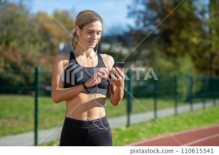 A fit female athlete in athletic wear selects music on her phone before a run in a sunny park setting. A fit female athlete in athletic wear selects music on her phone before a run in a sunny park setting. 110615341
