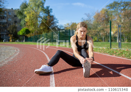 A determined woman stretches her leg muscles before a run on a track field, illustrating fitness and preparation. 110615348