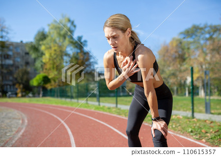 Athletic woman taking a break from a run on a sunny day, holding her knee and catching her breath on a track. 110615357