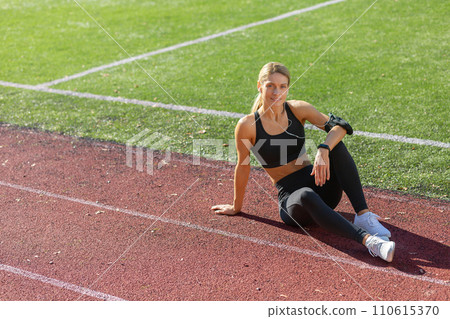Athletic woman wearing sportswear and earphones taking a break while sitting on a track field, enjoying a sunny day. Athletic woman wearing sportswear and earphones taking a break while sitting on a track field, enjoying a sunny day. 110615370