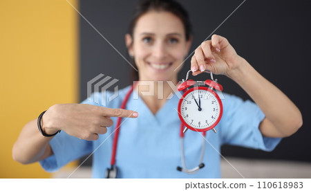 Smiling nurse points finger at red alarm clock Smiling nurse points finger at red alarm clock 110618983