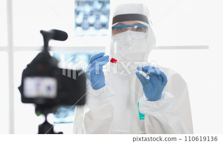 Doctor in protective suit demonstrates to camera test tube with shelf for sampling biological material Doctor in protective suit demonstrates to camera test tube with shelf for sampling biological material 110619136