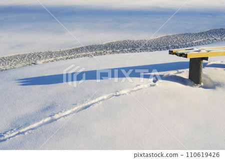 Frozen river. Winter wonderland. Snow-blanketed bench. Traces of wild animals in the snow. Winter specific. Frozen river. Winter wonderland. Snow-blanketed bench. Traces of wild animals in the snow. Winter specific. 110619426