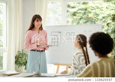 Cheerful UI designer answering questions of colleagues when speaking about mobile app interface in meeting Cheerful UI designer answering questions of colleagues when speaking about mobile app interface in meeting 110620597
