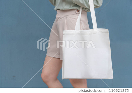 Blank white tote bag canvas fabric with handle mock up design. Close up of woman holding eco or reusable shopping bag against blue metal wall. No plastic bag and ecology concept. Blank white tote bag canvas fabric with handle mock up design. Close up of woman holding eco or reusable shopping bag against blue metal wall. No plastic bag and ecology concept. 110621275