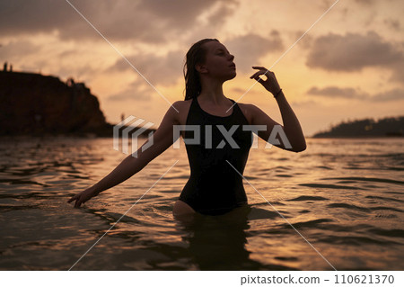 Silhouetted woman stands in sea at dusk, touching chin thoughtfully, serene horizon backdrop. Reflective swimmer enjoys evening calm, ocean expanse stretching behind. 110621370