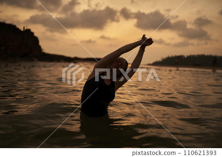 Serene dusk ocean swim. Woman stretches gracefully in calm seawater, silhouetted against amber sunset. 110621393