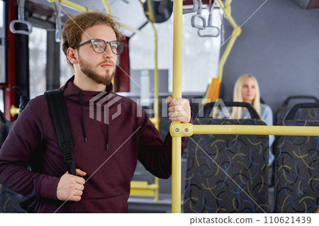 Close up of pensive adult male standing looking at window in public transport. Crop of cute young man in burgundy hoodie travelling with blur blonde sitting in urban transport. Concept of city life. Close up of pensive adult male standing looking at window in public transport. Crop of cute young man in burgundy hoodie travelling with blur blonde sitting in urban transport. Concept of city life. 110621439