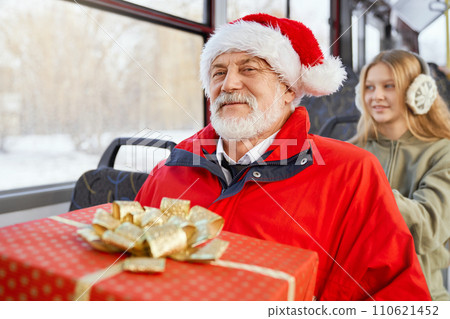 Front view of mature bearded male in red jacket smiling at camera and young girl sitting behind in earmuffs in public transport. Crop of passengers travelling by bus on holydays. Concept of joy. 110621452