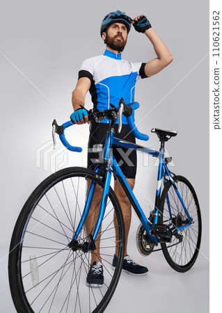 Strong motivated male cyclist getting ready to start in studio. Low angle view of bearded biker with blue bike looking away, while wearing helmet, isolated on grey. Concept of cycling, sport activity. 110621562