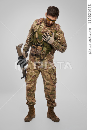 Weak soldier in camouflage outfit covering wound with hand indoors. Front view of wounded bearded military man checking chest, is there an injury, on gray background. Injury, rescue concept. Weak soldier in camouflage outfit covering wound with hand indoors. Front view of wounded bearded military man checking chest, is there an injury, on gray background. Injury, rescue concept. 110621658