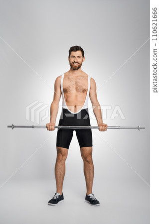 Strong bearded powerlifter posing with unweighted barbell in studio. Front view of smiling male bodybuilder in singlet looking at camera, on white background. Concept of powerlifting, sport. 110621666