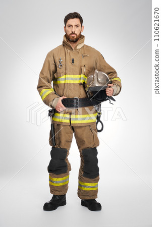 Serious handsome caucasian firefighter in full gear looking at camera indoors. Front view of strong male rescuer in uniform holding protective helmet, on white studio background. Workplace concept. Serious handsome caucasian firefighter in full gear looking at camera indoors. Front view of strong male rescuer in uniform holding protective helmet, on white studio background. Workplace concept. 110621670