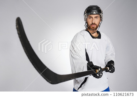 Bearded hockey player in protective shield pointing black stick into camera. Close up view of caucasian male ice hockey sportsman in uniform performing slap shot, isolated on gray. Concept of sport. Bearded hockey player in protective shield pointing black stick into camera. Close up view of caucasian male ice hockey sportsman in uniform performing slap shot, isolated on gray. Concept of sport. 110621673