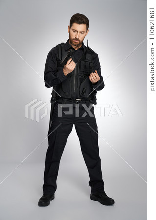 Collected caucasian police officer reloading weapon, getting ready to shoot. Front view of man in cop uniform recharging handgun, holding magazine, on gray studio background. Work, weapon concept. Collected caucasian police officer reloading weapon, getting ready to shoot. Front view of man in cop uniform recharging handgun, holding magazine, on gray studio background. Work, weapon concept. 110621681