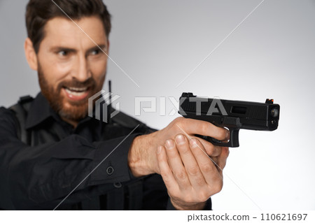 Persistent dark-haired policeman keeping gun both hands, aiming indoors. Close up of black firearm, pistol, held by blurred silhouette of aggressive cop, on gray background. Concept of work, weapon. 110621697