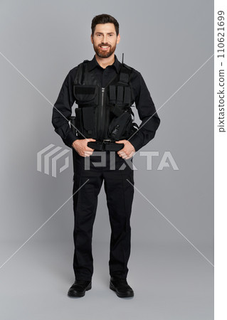 Happy police officer with dark hair posing, with hands on belt in studio. Front view of attractive cop in full gear smiling to camera, on gray background. Concept of work, profession, police. 110621699