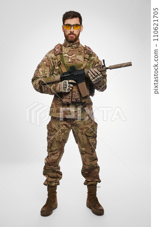 Confident, serious military officer carrying assault weapon with both hands in studio. Front view of bearded infantryman in camouflage, posing with shotgun, on white background. Concept of army. 110621705