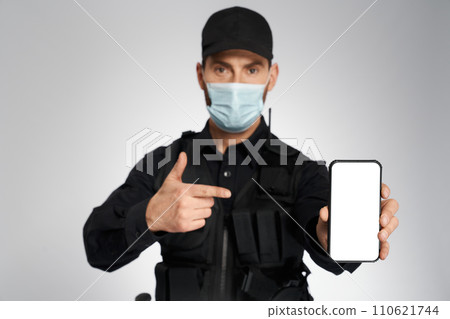 Male policeman in protective mask pointing to empty cell phone's screen. Portrait of caucasian cop gesturing, while looking camera, on gray studio background. Concept of safety, calling, quarantine. Male policeman in protective mask pointing to empty cell phone's screen. Portrait of caucasian cop gesturing, while looking camera, on gray studio background. Concept of safety, calling, quarantine. 110621744