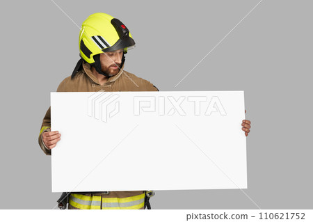 Focused male firefighter looking at blank banner in front. Front view of fireman in helmet holding big white cardboard with copy place, isolated on gray studio background. Workplace, advert concept. 110621752
