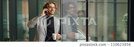 Cheerful businessman in eyeglasses is talking phone standing in modern office near window Cheerful businessman in eyeglasses is talking phone standing in modern office near window 110623856